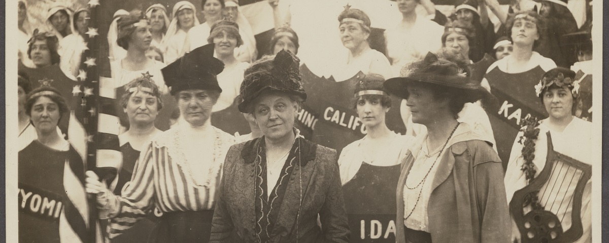 Carrie Chapman Catt stares defiantly at the camera. She is surrounded by women protesting for their right to vote. Some carry shields with state names on them, California, Wyoming, Kansas. Others carry American flags.