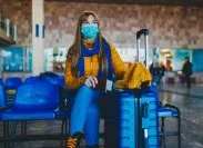 A woman wearing a face mask sits next to her blue suitcase in a travel lounge